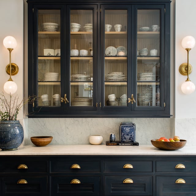 A transitional kitchen features dark blue glass-front upper cabinets and matching base cabinets with brass hardware, flanking a white marble backsplash and countertop decorated with bowls, fruit, and a blue and white jar.